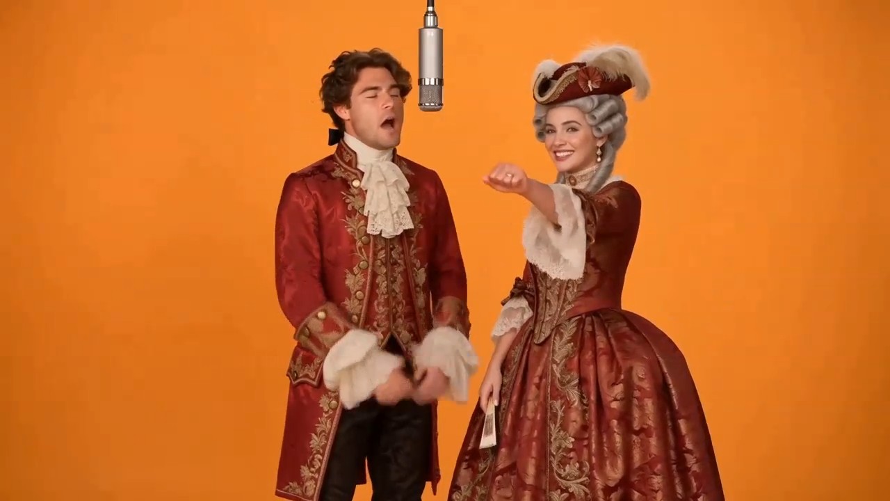 An 18th-century rococo duet: a man in a burgundy brocade frock coat and a woman in a pannier gown with powdered wig sing playfully into a hanging studio microphone against a bright orange backdrop.